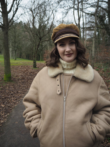A woman wearing a shearling aviator jacket and cord baker boy hat stands on a winter treelined path in a park