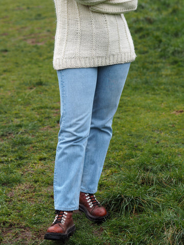 A woman stands in a field with her arms crossed wearing a cream wool jumper, a pair of blue Levis 501 jeans and a pair of chestnut brown hiking boots