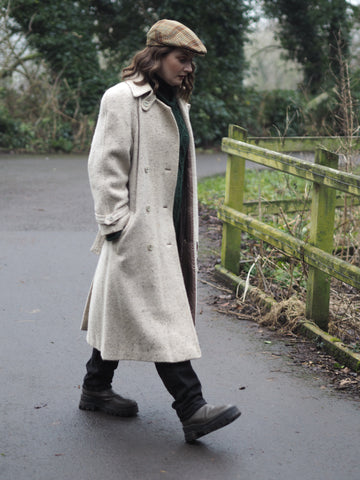 A woman walks along a river towpath wearing a cream flecked tweed trench coat, a green aran knit sweater, a wool check flat cap, black jeans and a pair of green tractor sole boots