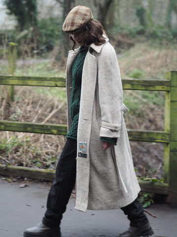 A woman walks along a river towpath wearing a cream flecked tweed trench coat, a green aran sweater, black jeans, a wool check flat cap and a pair of green tractor sole boots