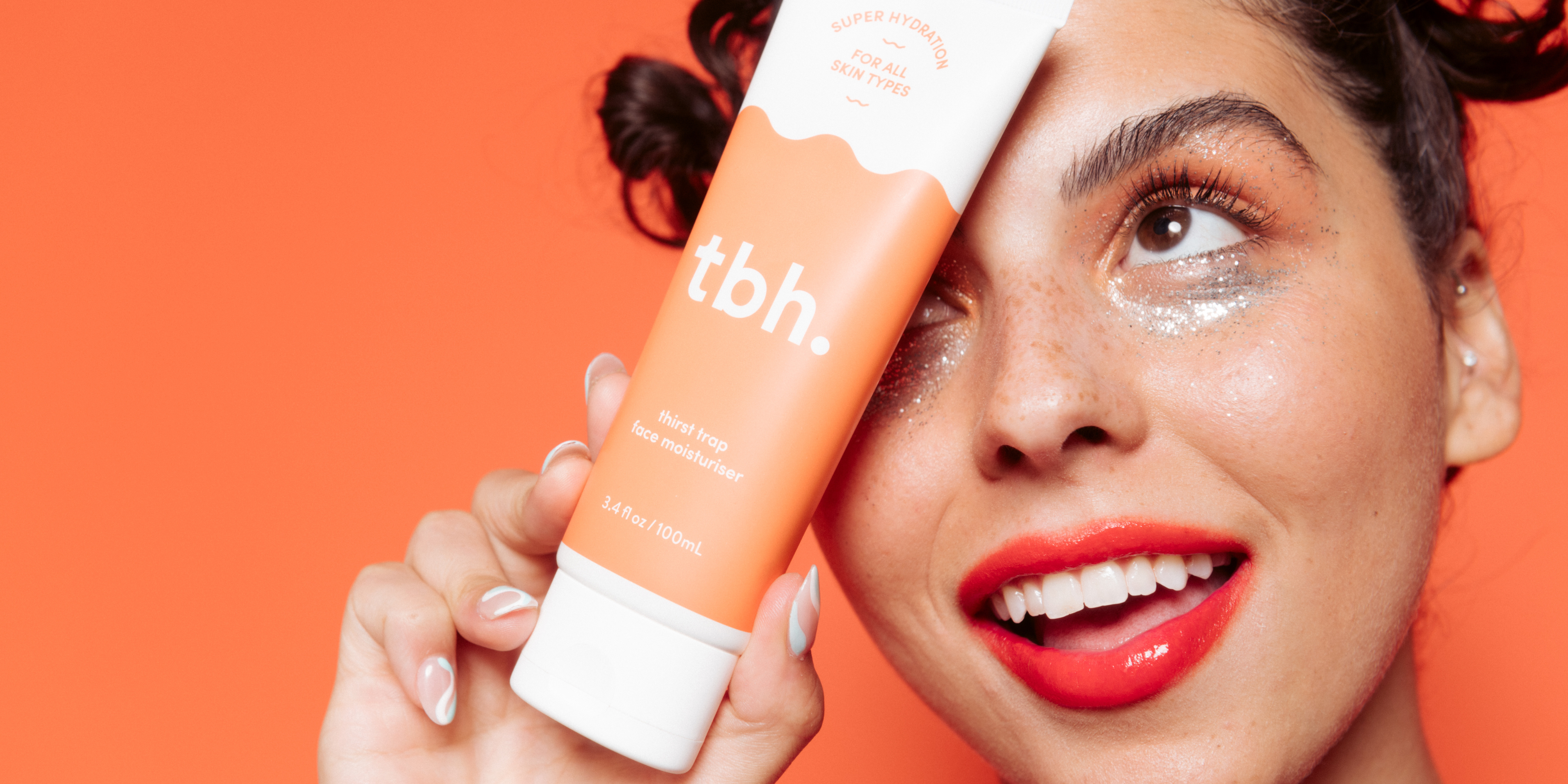 Orange background, girl with pink eyeshadow and glitter under her eyes, red lipstick smiling and lookinbg up with tbh skincare thirst trap moisturiser. Dry skin ingredients