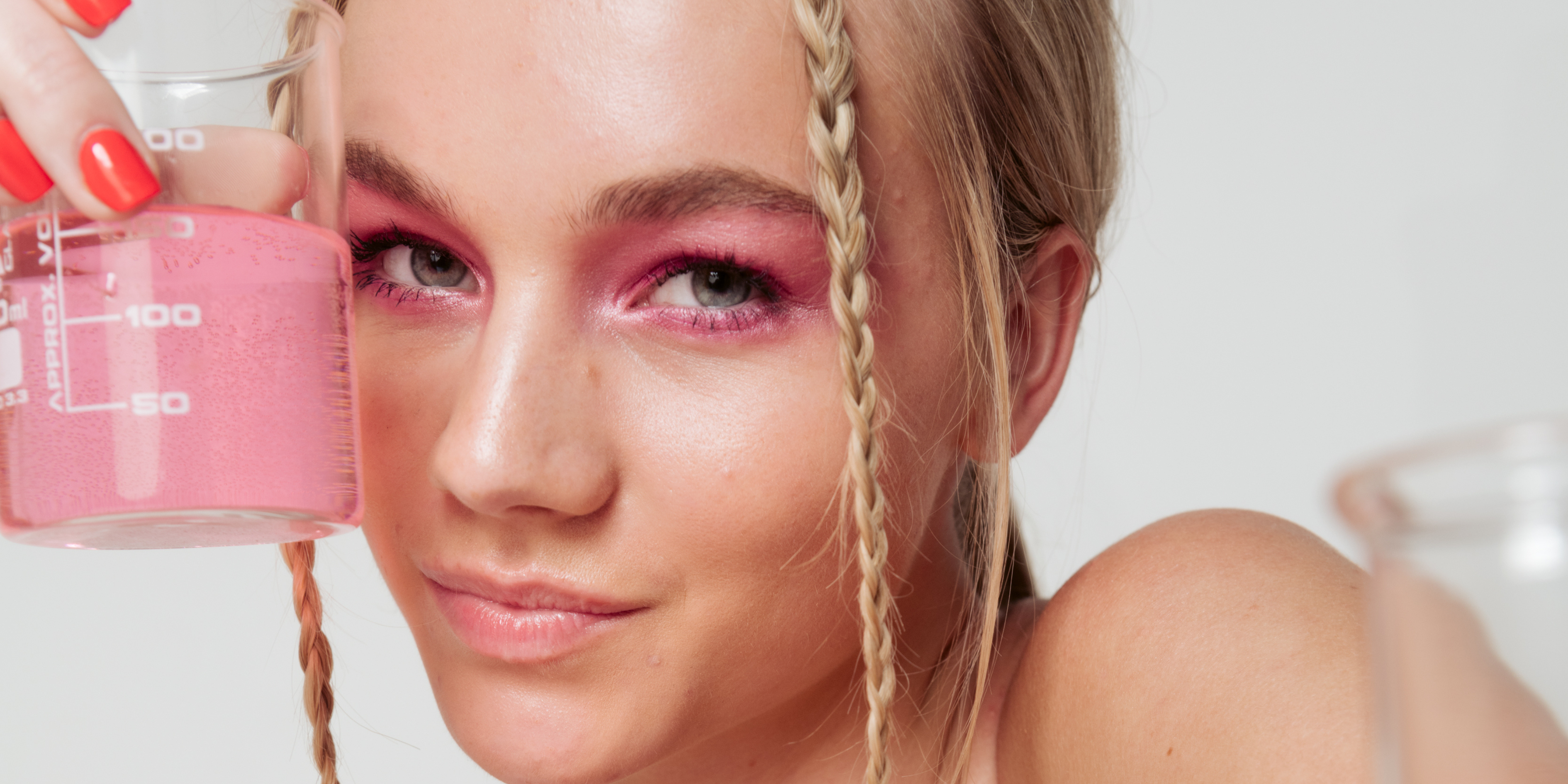 girl with blonde hair and pink eye shadow, holds beaker full of pink liquid to her face