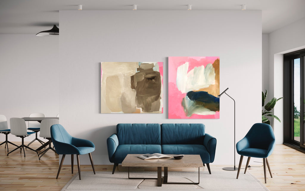 A sophisticated living room space featuring two large abstract paintings on a white wall, complemented by modern furniture. On the left, a painting with neutral tones of beige and brown, and on the right, a vibrant piece with pink, cream, navy, and touches of gold. The room is furnished with a plush teal sofa, two matching armchairs, and a minimalist coffee table, all set upon light wooden flooring, creating a harmonious blend of contemporary art and stylish interior design.