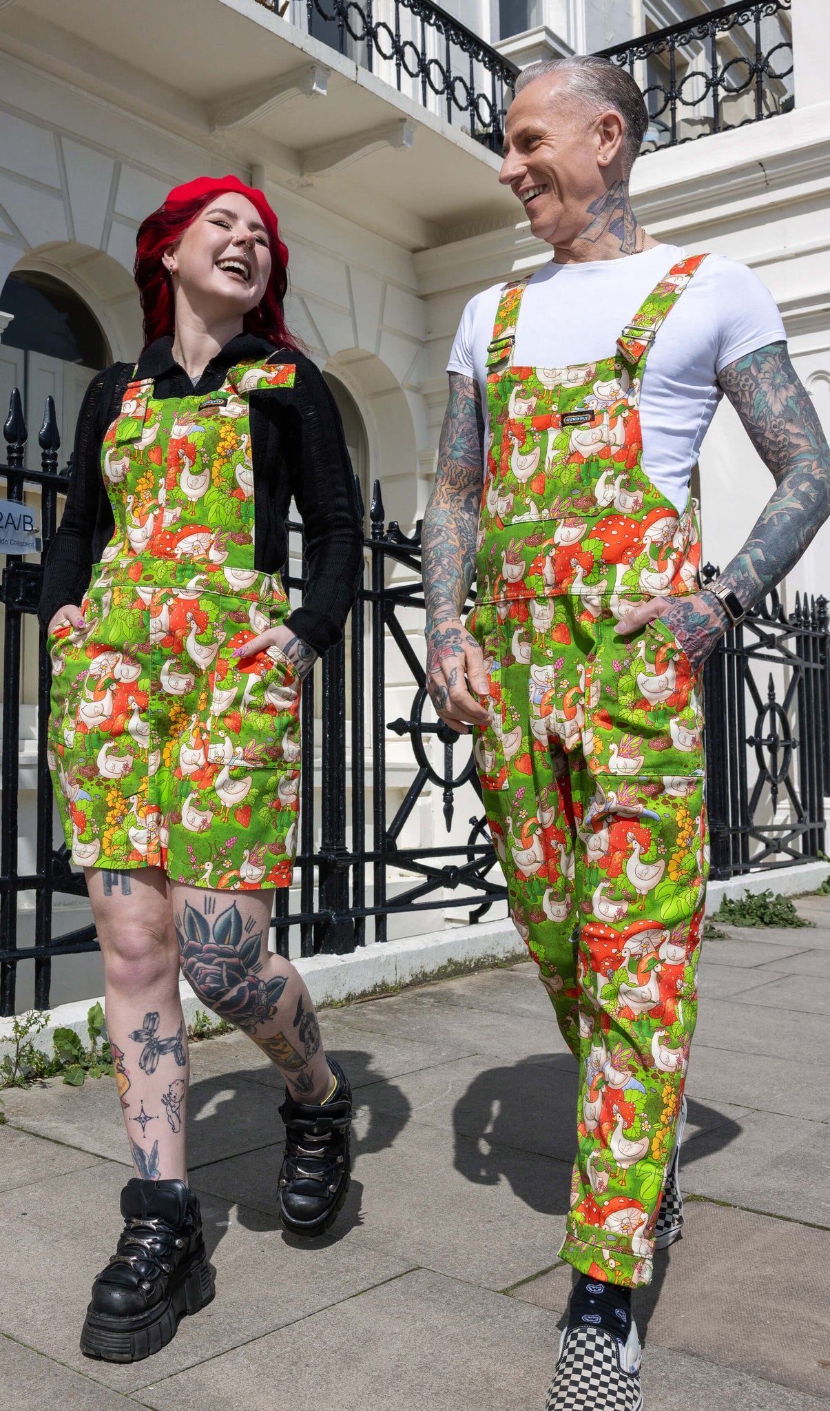 The Run & Fly x The Mushroom Babes In The Geese Garden Stretch Twill Pinafore Dress being worn by Flo with a long sleeve black top, black trainers and red mushroom beret. She has red and black hair and tattoos, she is walking down a pavement laughing with James wearing the dungarees. The print features geese wearing various cute hats, such as mushrooms and frogs, collecting flowers and strawberries on green grass.