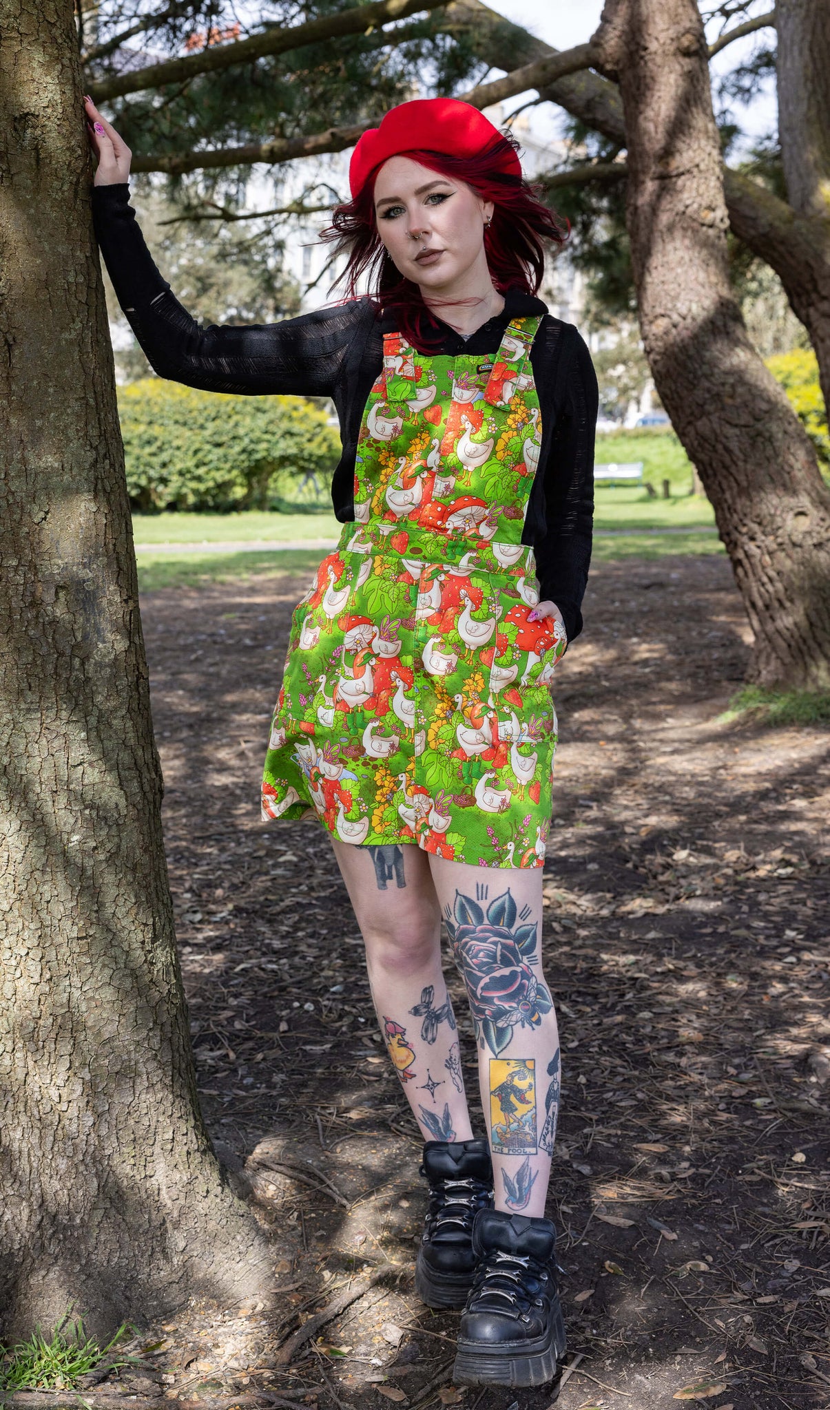 The Run & Fly x The Mushroom Babes In The Geese Garden Stretch Twill Pinafore Dress being worn by Flo outside in a park with a long sleeve black top, black trainers and red mushroom beret. She has red and black hair and tattoos, she is facing forward smising leaning one hand on a tree with the other in the dress pocket. The print features geese wearing various cute hats, such as mushrooms and frogs, collecting flowers and strawberries on green grass.