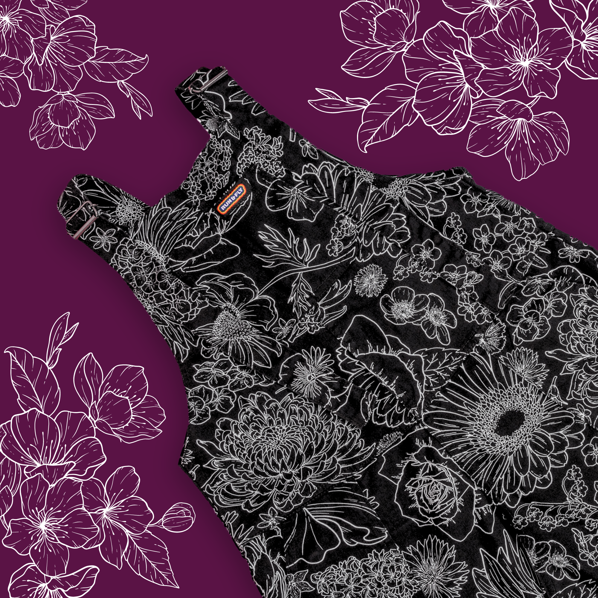 Black floral dungarees laid on a burgundy red background. The ethically made dungarees are a black base with white floral linework all over with retro roller straps and a rainbow run and fly tag sewn to the top of the front bib.