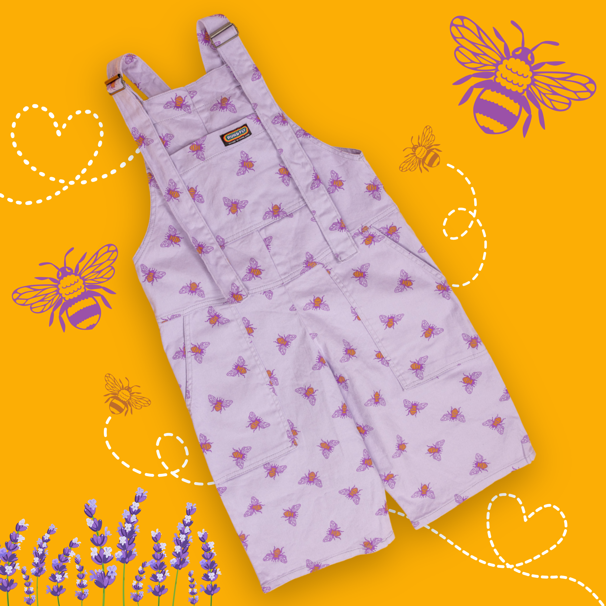 Flat lay of lavender base colour dungaree shorts with yellow buzzy bumble bees all over with the run and fly rainbow tag sewn on the top of the front bib. The background of the photo is yellow with trailing heart line following bees and lavender sprigs at the bottom.