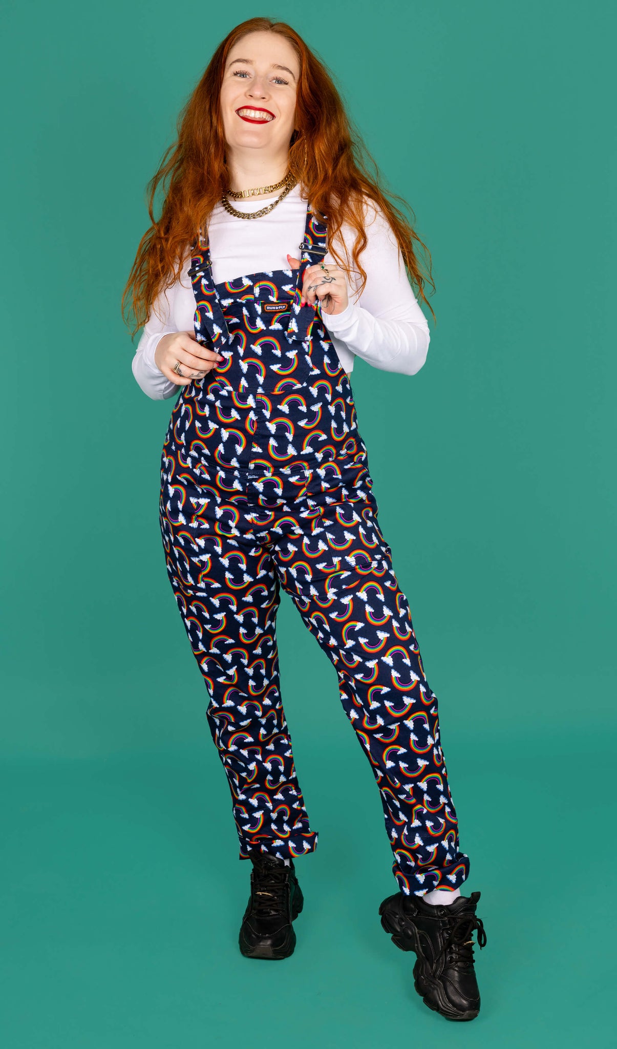 Isobella a white femme model with long ginger hair is wearing navy blue dungarees with all over rainbow and cloud print paired with long sleeve white top and black trainers. Isobella is smiling at the camera with one leg in front of the other and is holding the front of the dungarees in front of a green background.