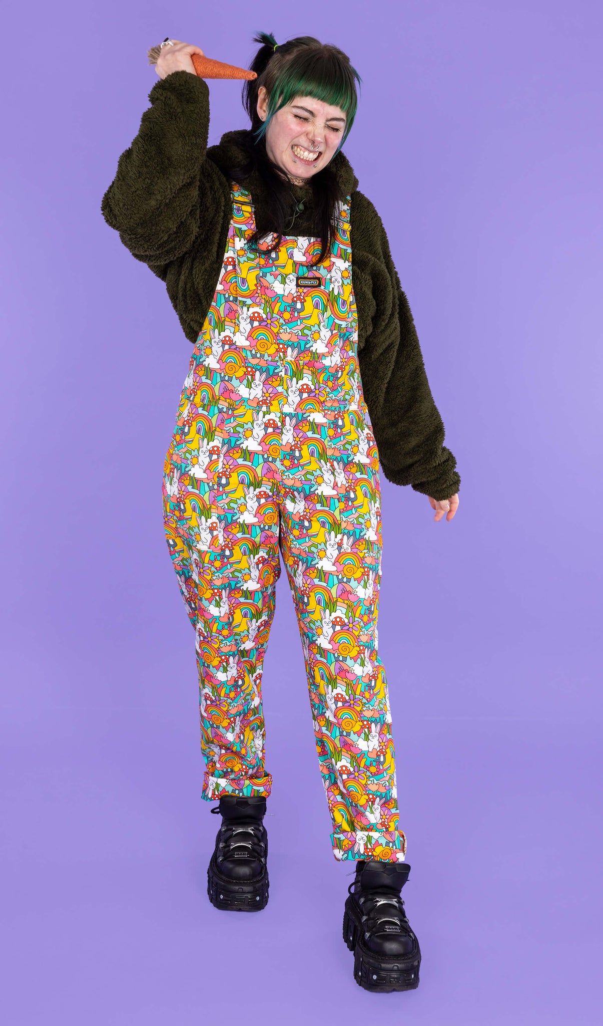 Faeryn wearing the some bunny loves you stretch twill dungarees from run and fly with a green fluffy jumper and black platform trainers. The print features rainbows, bunnies, mushrooms and smiling snails. They are facing forward on a purple background holding up a prop carrot with a scrunched up smiling face.