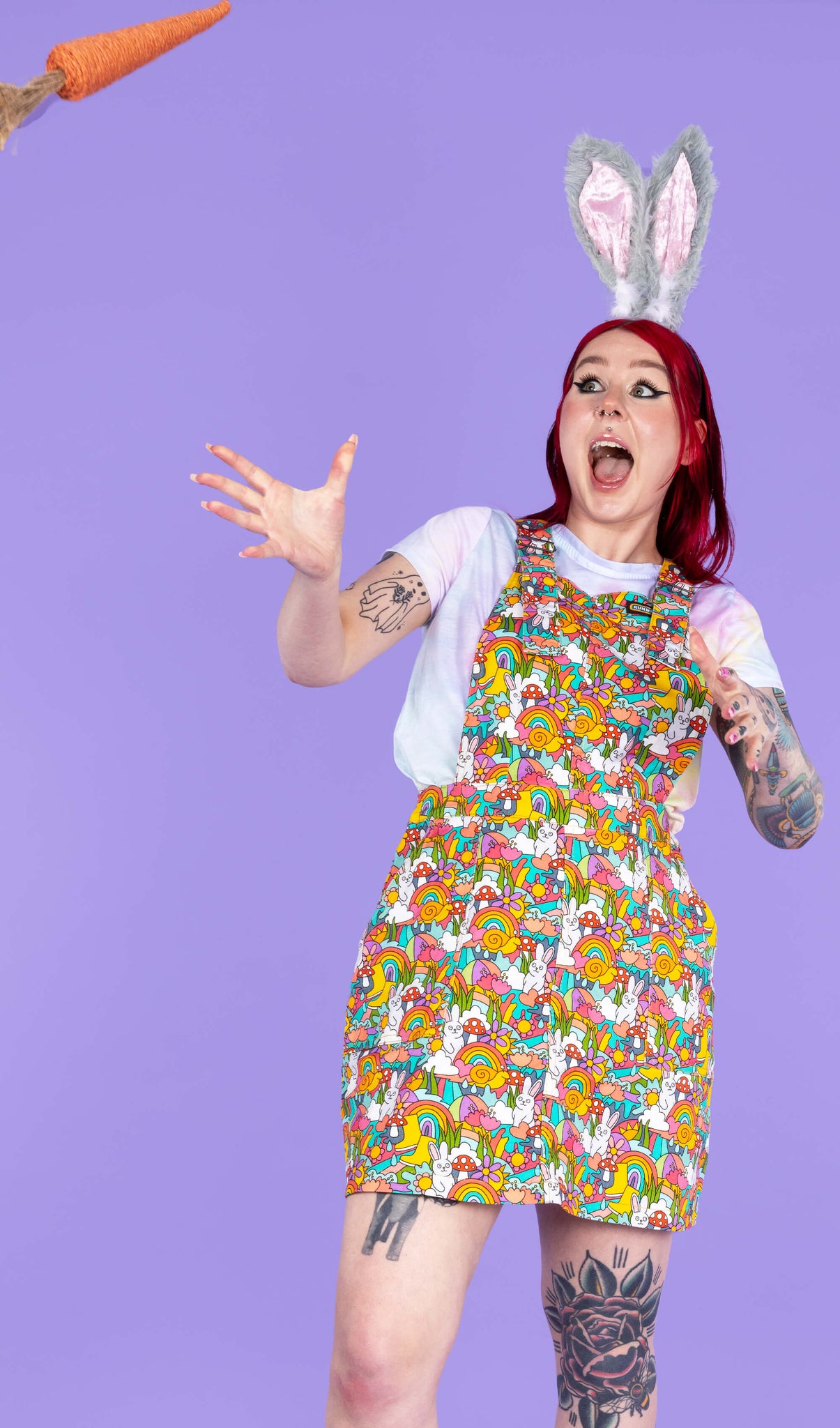 Flo modelling the some bunny loves you stretch twill pinafore by Run and Fly, the print features rainbows, bunnies, mushrooms and smiling snails all over. She is stood facing forward on a purple background with a shocked expression as a prop carrot flies towards her.