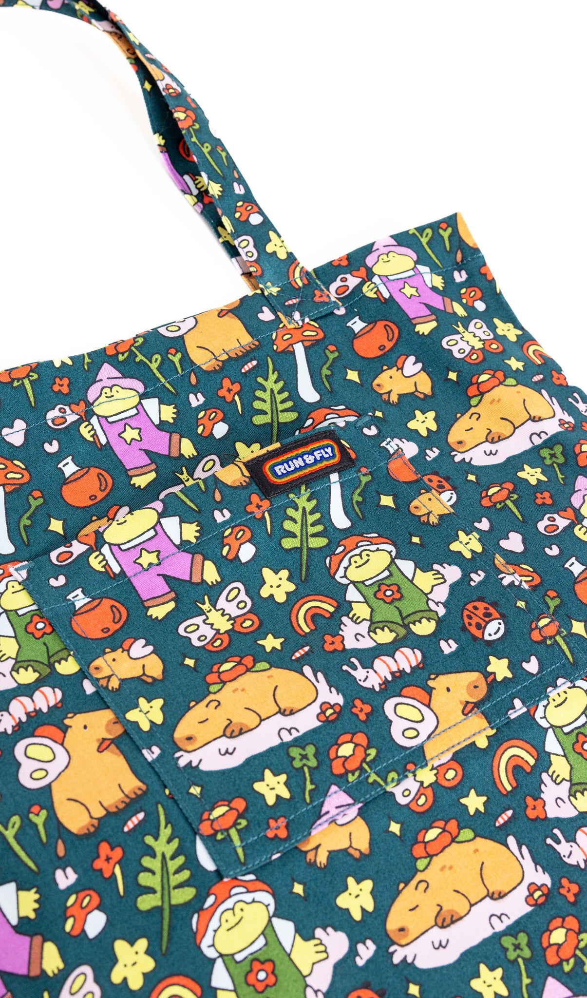 The Run & Fly x Nimbob Capybara Fairies Tote Bag laying flat on a white background. The green base tote bag feature frogs in dungarees, butterflies, plants, potion bottles, mushrooms and capybaras with fairy wings.