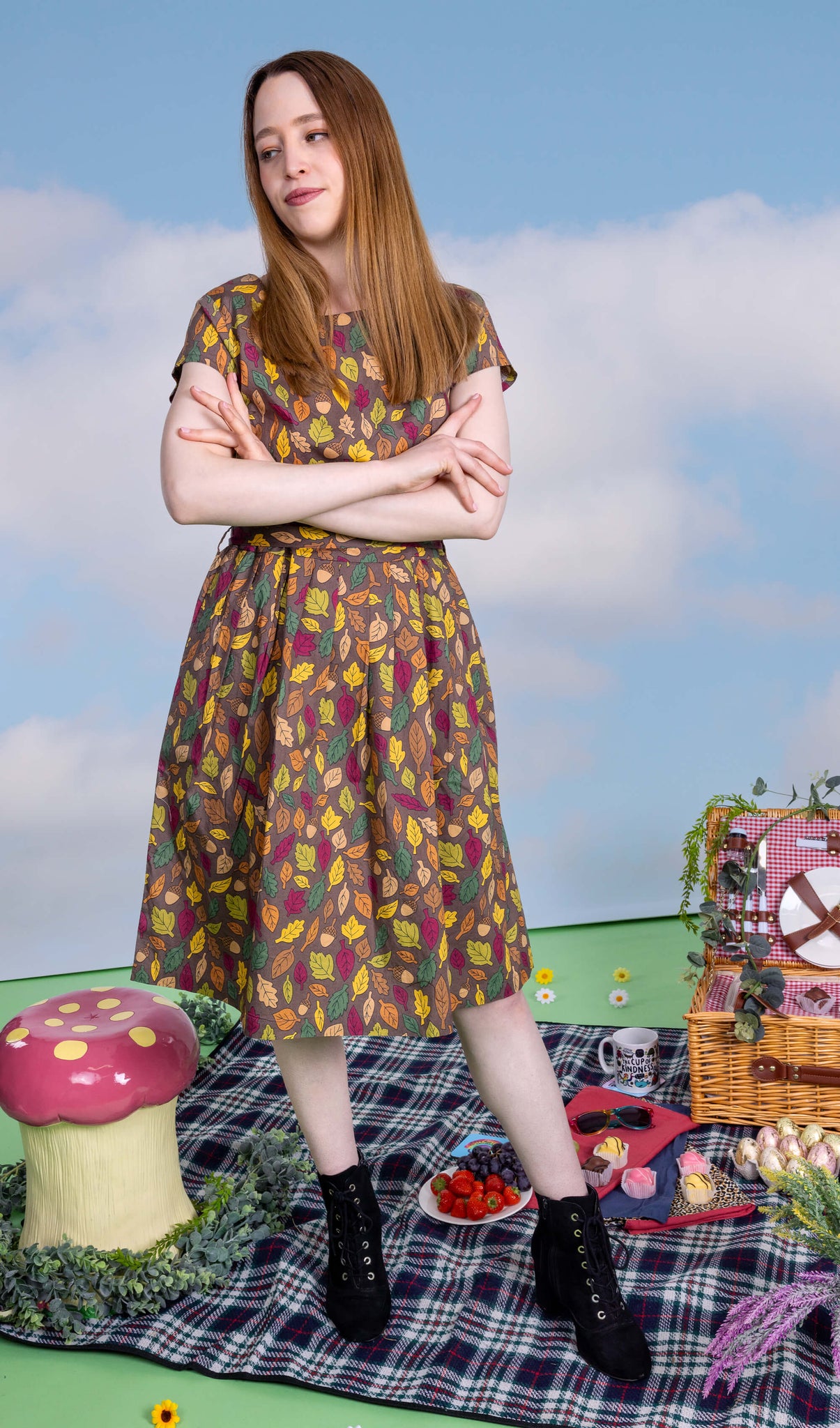 The Autumn Leaves Stretch Belted Tea Dress with Pockets on a femme model with long brown hair and black heeled boots. She is posing leaning back on one leg with her arms crossed smiling to the left at a picnic setup with a picnic basket, fruit, flowers and katie abey mugs. The dress print is a brown base with green, red, orange and yellow autumnal leaves and acorns.