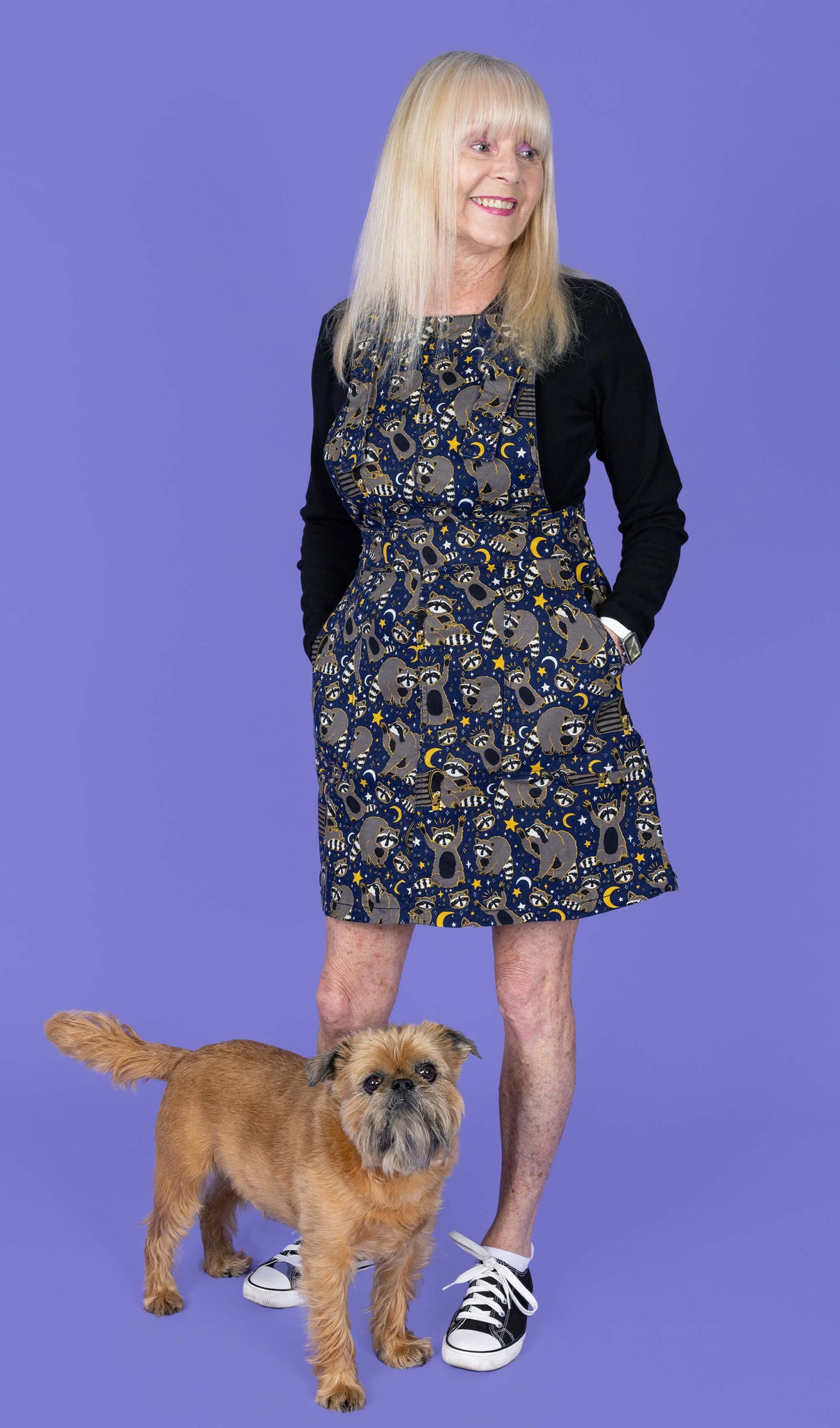 The Trash Pandas Stretch Twill Pinafore Dress worn by a femme model with blonde hair, a black long sleeve tshirt and black converse style trainers on a purple studio background with a small brown dog at her feet. She is facing forward smiling looking off to the right with both hands resting in the side pockets. The print is a rich dark blue with grey racoons, trash bins, white and yellow sparkles, yellow stars and moons and small eyes.