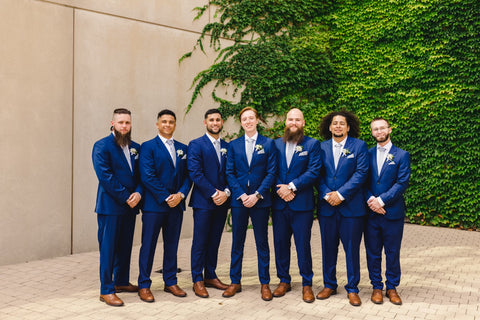 Seven young men, all wearing blue suits, standing, smiling in front of a beige wall with trees on the right.