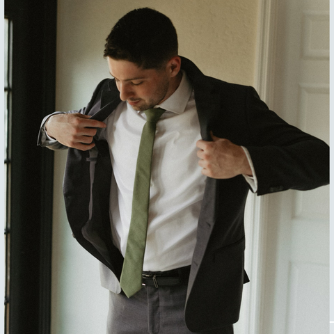Man with dark hair putting on his suit jacket. The suit is grey and he is wearing a white button down shirt with a moss green tie.