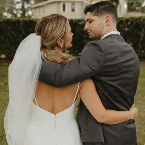 We see the back of a bride and groom, waist up. The bride is on the left, the groom on the right. They are looking directly at each other so we see their profiles. They have their arms around one another.