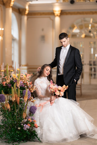 Man looking down at bride, who is sitting holding a bouquet of flowers.