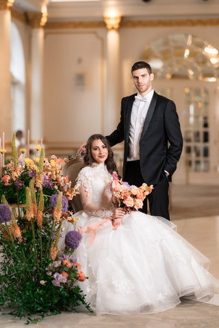 Bride sitting holding bouquet of flowers with grom standing by her side.