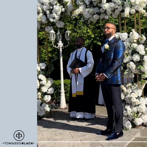 Man in blue and black patterned tuxedo stnading on alter outside with white flowers in the background.