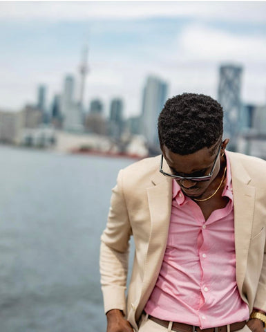 close up of young man, wearing sunglasses, a beige suit, and a pink button down shirt, no tie.