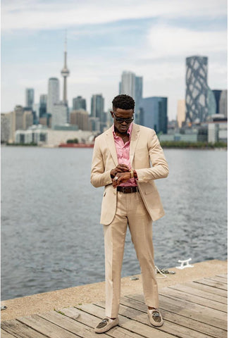 Full length image of man in beige suit, with a pink button down, standing on a pier, holding his hands together.