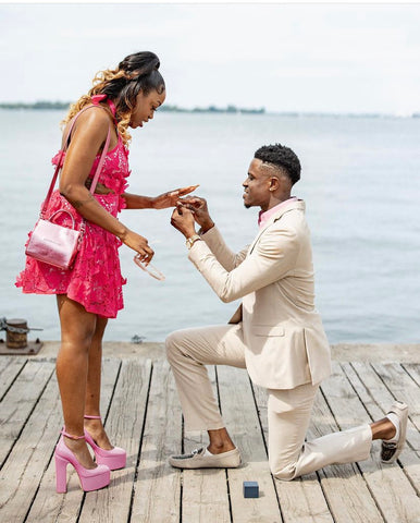 Man, on right, on one knee proposing to a woman, on the left. He is wearing a beige suit, and she is wearing a bright pink dress. 