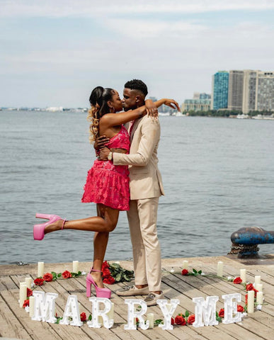 Man and woman hugging. They are standing on a pier, the water is behind them. On the floor surrounding them are the words "marry me" with roses.