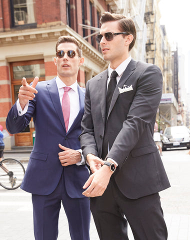 Two men wearing sunglasses, one in a dark grey suit, the other in blue. Both looking the same direction, one is pointing at something but we don't know what it is.