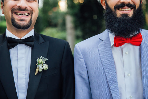 Close up of two men smiling; one wearing a balc suit with peak collar and black bow tie, the other wearing a light blue suit with notch collar and red bow tie