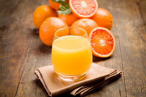 Image shows a bunch of oranges in the background and a glass of freshly-squeezed orange juice in the foreground