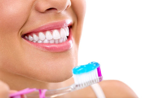 Close-up of a smiling young woman's mouth full of a healthy teeth. Image also shows a toothbrush loaded with blue toothpaste in the foreground
