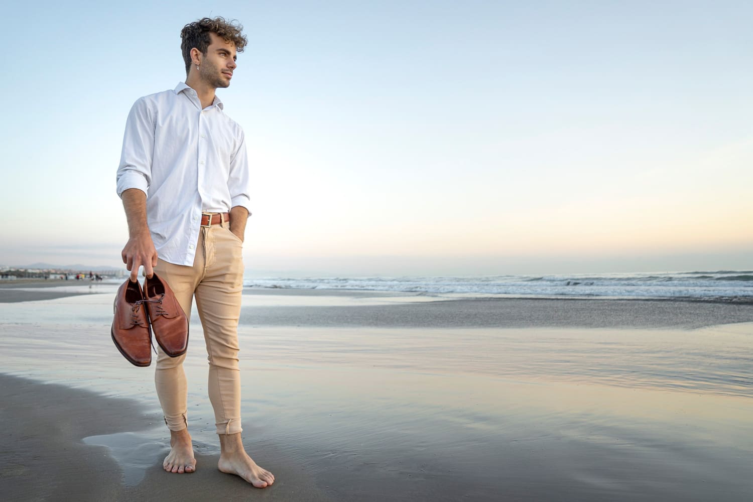 Semi-Formal Beach Wedding Attire image, Classy elegant man in white shirt and beige pants walking on the beach with his shoes in hand