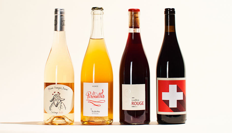 A line-up of four wine bottles. From left to right; a light rose with white label, an orange-colored wine with a crown cap, a red wine with its cork visible through the clear glass wine bottle, and a red wine with a red label.