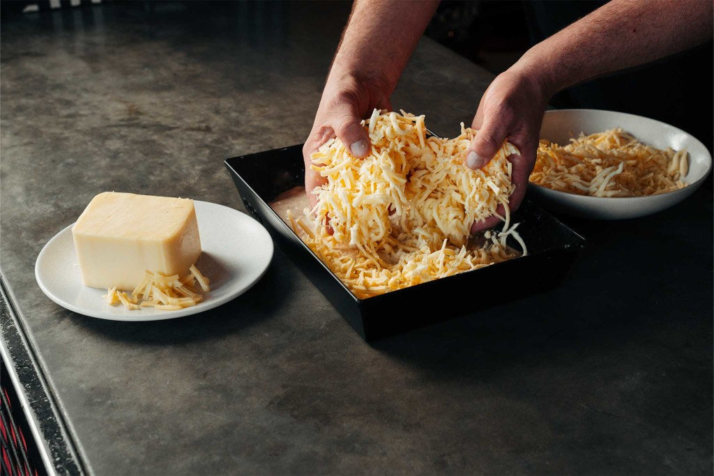 Two hands placing shredded mozzarella and cheddar over dough in an Ooni Detroit-style Pizza Pan on a table next to a dish of shredded cheese and a plate of cheddar.
