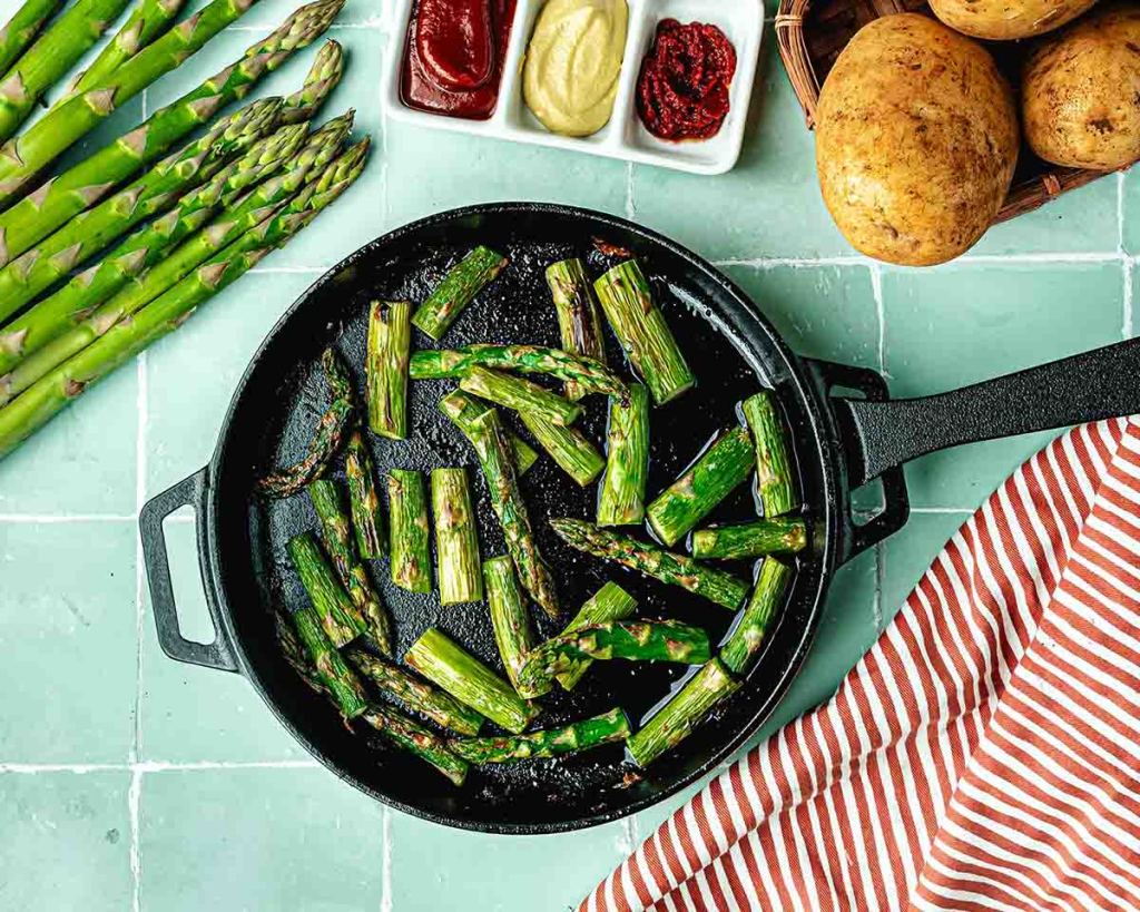 Cooked pieces of asparagus in an Ooni Cast Iron Skillet Pan on a table next to asparagus, potatoes and a dish of condiments.
