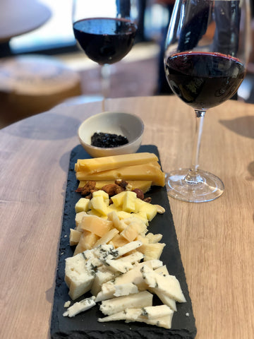 Introduction picture for the best cheese boards in leeds city centre, showing a black slate board covered in a heap of cheese, with two glasses of red wine behind it