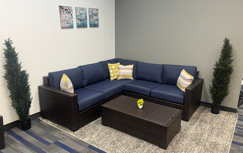 Brown hand-woven wicker sofa and coffee table, with white throw pillows.  Small trees on both sides.