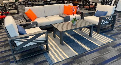 Modern grey metal aluminum sofa set with orange throw pillows, 2 club chairs, and coffee table with an orchid on it.