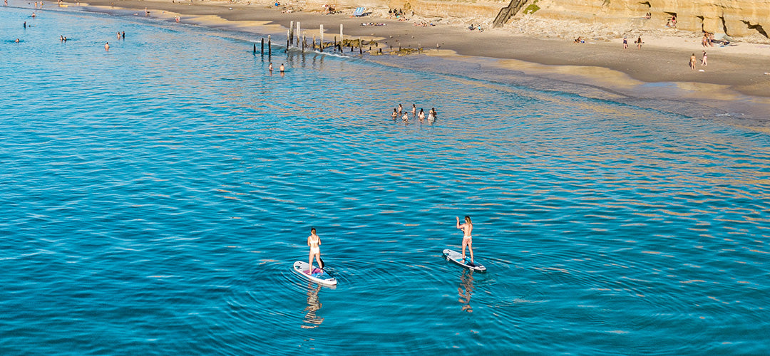 A man and woman stand up paddleboarding with other people swimming around