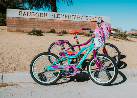 Two different colored Elm 18 kids bikes by Schwinn pictured outside of the Sanborn Elementary School sign