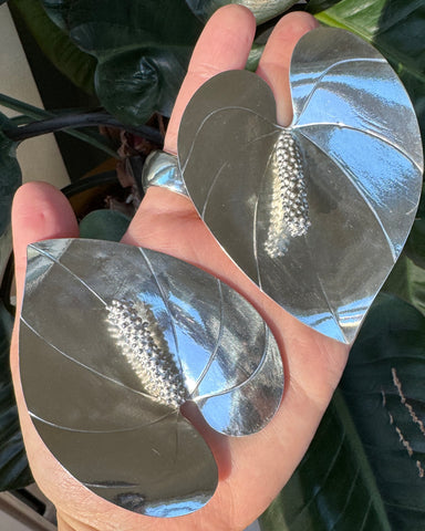 photo of handmade silver anthurium flower earrings pointing in different directions, displayed laying flat in the palm of a hand with green plant leaves in the background