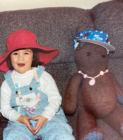 A photo of the author as a child dressed in overalls and a pink hat sitting with her brown bear friend on a grey couch, both wearing handmade beaded pastel necklaces