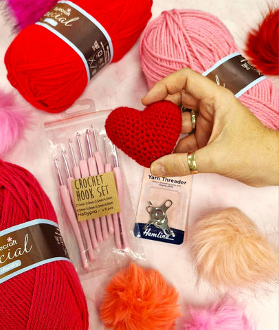 Flatlay of pink and red balls of wool and crocheting accessories. In front a hand holds a crocheted heart