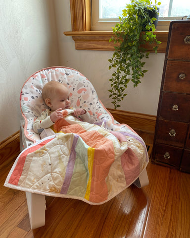 Baby in a swing snuggled in a small rainbow pastel colored quilt