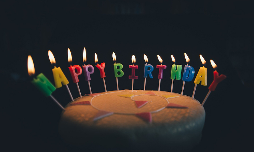 A birthday cake in a darkened room with lit candles shaped like letters that spell "happy birthday."