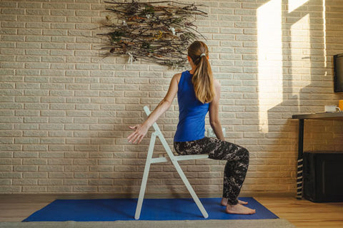 Woman doing yoga at home. Blue mat and undershirt. Twisting pose with a chair, sunny day. Natural panels on the brick wall, sustainable design. Attending to mental health.