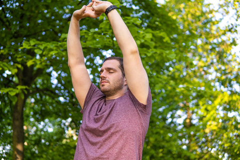 Photo of a young and attractive man in the park streching with his arms up to the sky and his eyes closed. Alone in nature during a summer day. Yoga pose