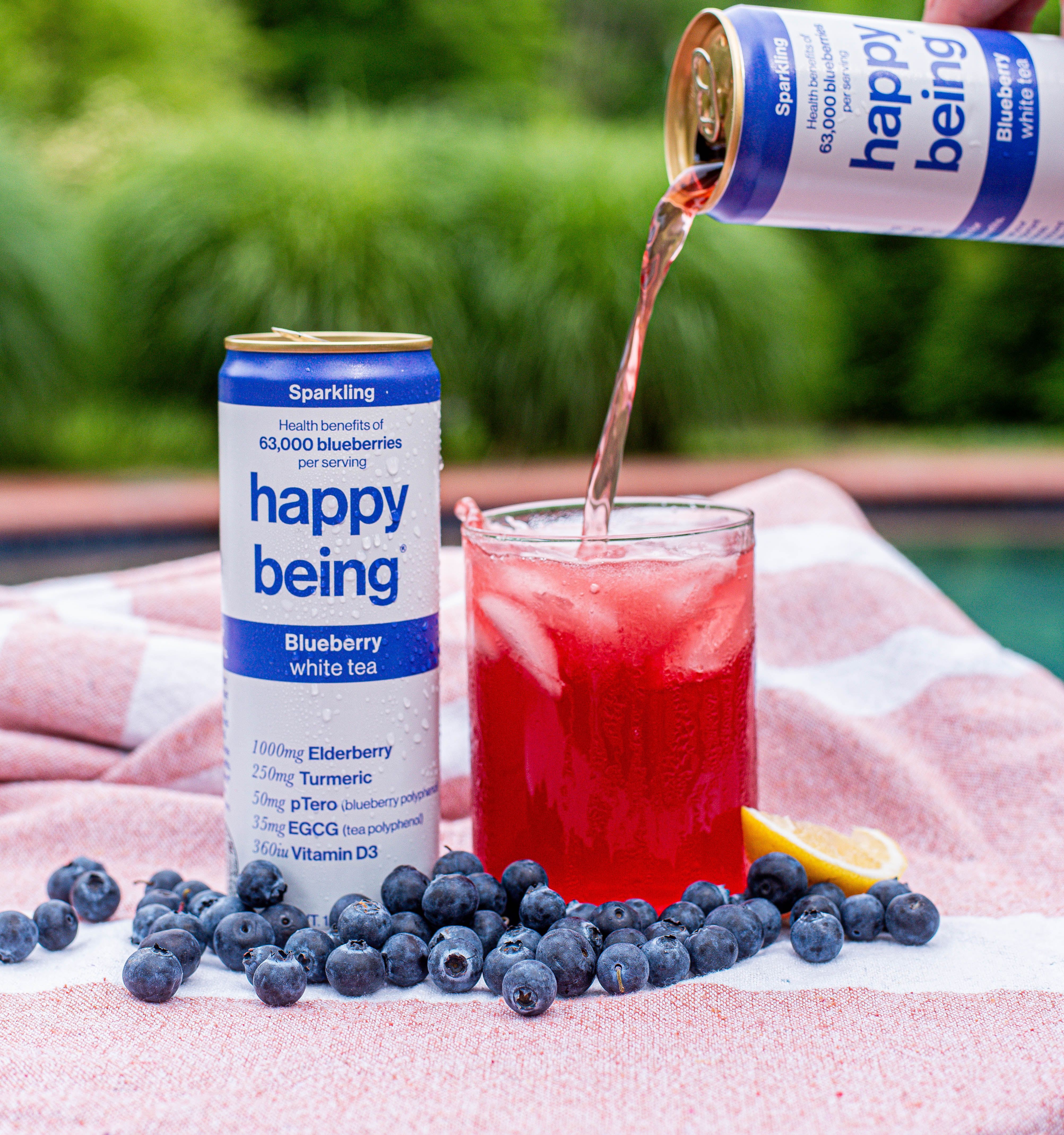 A can of sparkling blueberry white tea poured into a glass, surrounded by fresh blueberries and a lemon slice.