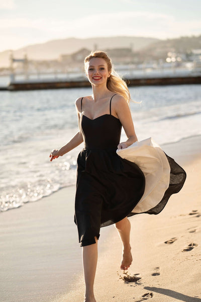 A blonde girl recreating Brigitte Bardot’s iconic run on the beach in Cannes, wearing a Gaâla cotton/linen black Bardot dress with petticoat