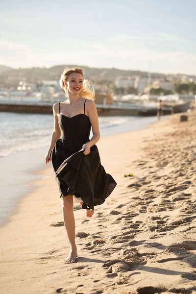 A laughing long haired blonde running down the sandy beach in Cannes, wearing Gaâla’s black cotton/linen Bardot dress